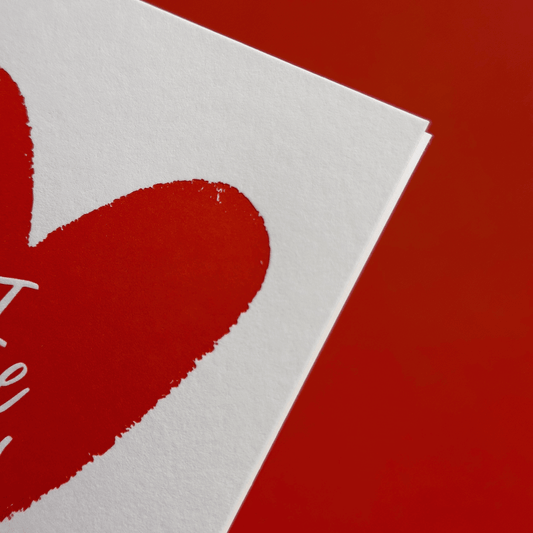 Closeup of red heart illustration on a white paper with 'I love you' hand lettering, against a red background