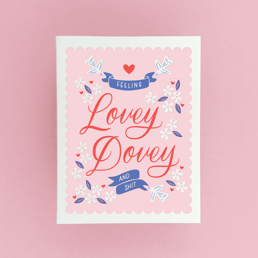 Letterpress love card displayed on light pink background with 'Feeling Lovey Dovey and Shit' in playful red and blue lettering.
