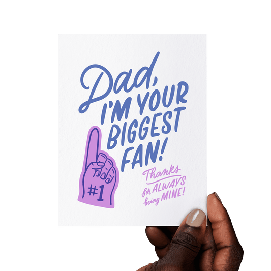 Hand holding a heartfelt Father’s Day card with ‘Dad, I’m your biggest fan! Thanks for always being mine!’ in blue and lilac hand lettering, with a foam finger illustration.