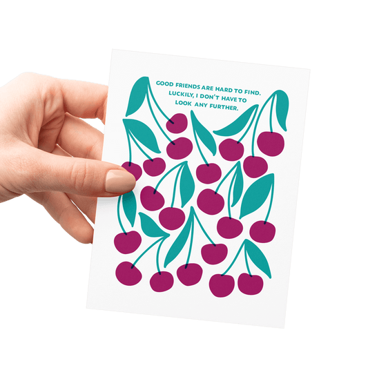 Hand holding a friendship card with ‘Good friends are hard to find. Luckily, I don’t have to look any further.’ in teal hand lettering, surrounded by merlot-colored cherries and green leaves.