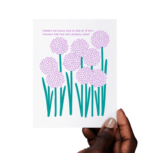 Hand holding a floral mom card with ‘There’s no place like in bed by 9 PM—thanks for the life lessons, Mom!’ in purple, surrounded by blooming alliums.