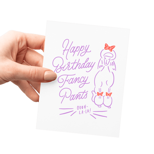 Hand holding a funny birthday card with 'Happy Birthday Fancy Pants' in purple hand lettering, featuring a poodle illustration with bows.