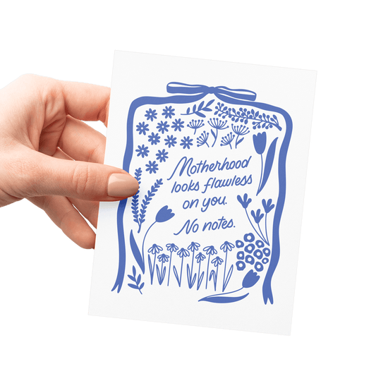 Hand holding a floral greeting card with ‘Motherhood looks flawless on you. No notes.’ in blue hand lettering, framed by vintage-inspired folk floral artwork.