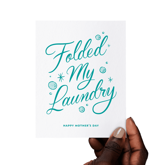 Hand holding a funny Mother’s Day card with ‘Folded My Laundry’ in teal hand lettering, accented with vintage-style bubbles and stars.