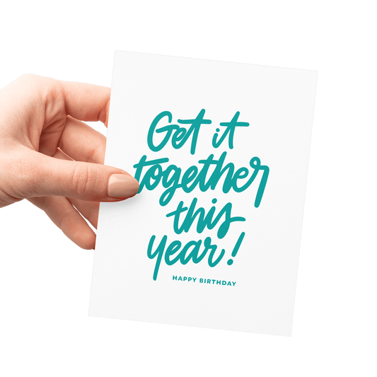 Hand holding a humorous birthday card with 'Get It Together This Year!' in teal hand lettering.