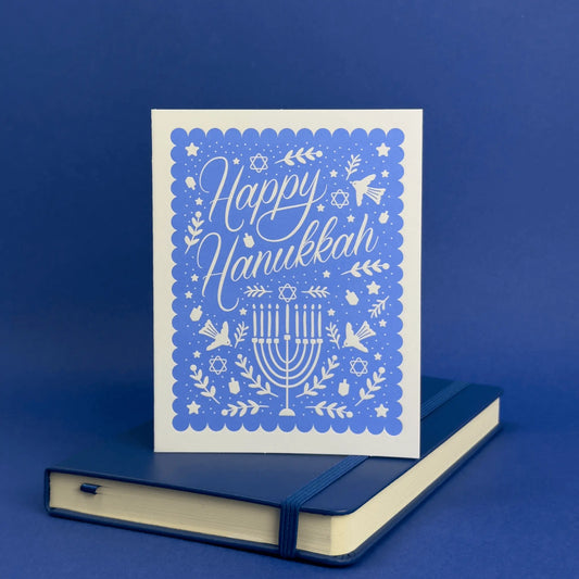 Card with 'Happy Hanukkah' text and menorah design on a blue background, placed on a matching blue notebook.