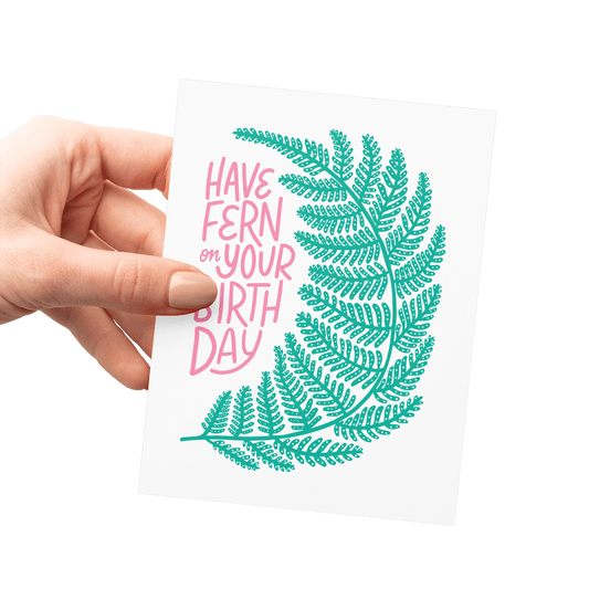 Hand holding a botanical birthday card with a detailed fern illustration and pink hand-lettered text.