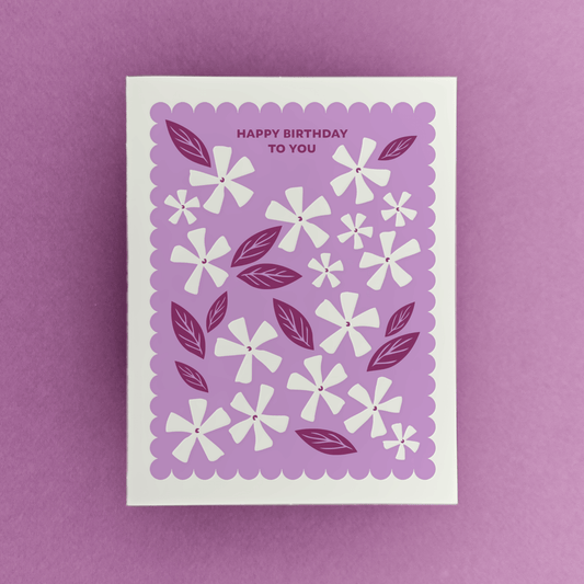 Letterpress birthday card with white floral illustrations, berry-toned leaves, and a purple scalloped border reading “Happy Birthday to You.