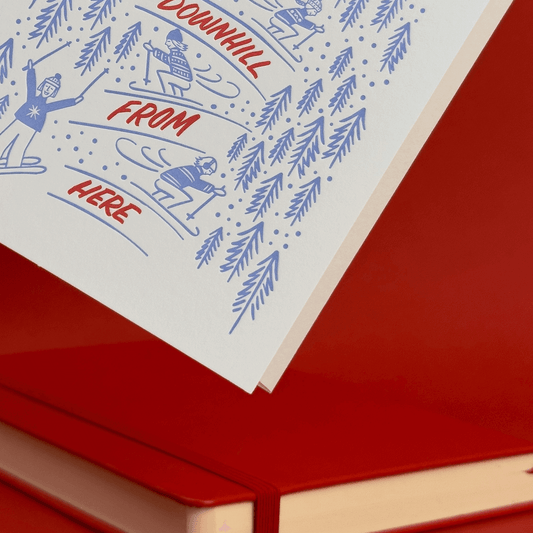 Skiing-themed greeting card with 'Downhill from here' hand lettering and ski illustrations on a red background