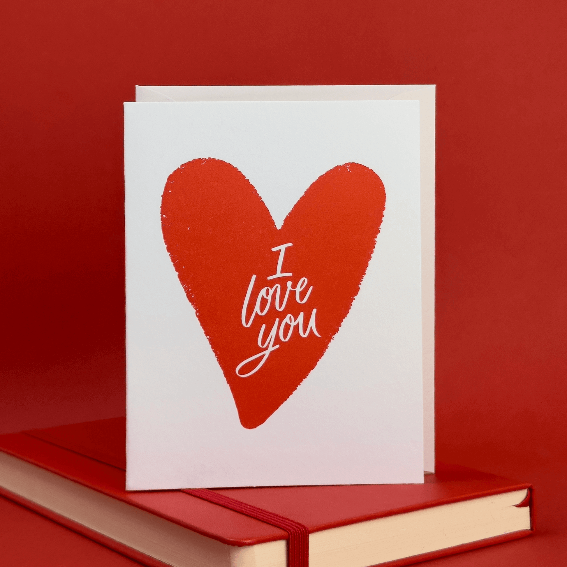 Card with a red heart illustration with 'I love you' hand lettered inside of it on a red background.