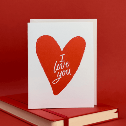 Card with a red heart illustration with 'I love you' hand lettered inside of it on a red background.