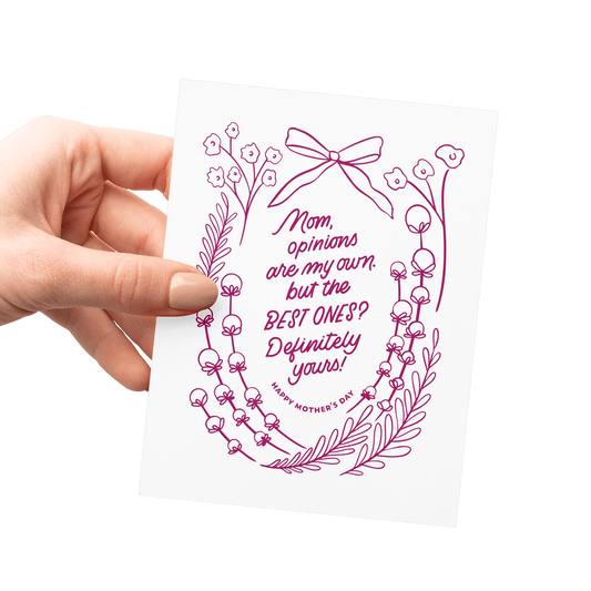 Hand holding a floral Mother’s Day card with ‘Mom, opinions are my own, but the best ones? Definitely yours!’ in burgundy hand lettering, surrounded by vintage floral artwork and a ribbon illustration.