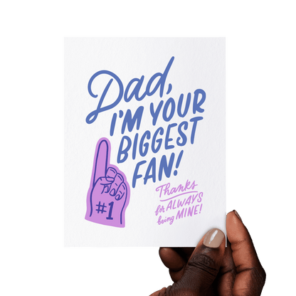 Hand holding a heartfelt Father’s Day card with ‘Dad, I’m your biggest fan! Thanks for always being mine!’ in blue and lilac hand lettering, with a foam finger illustration.