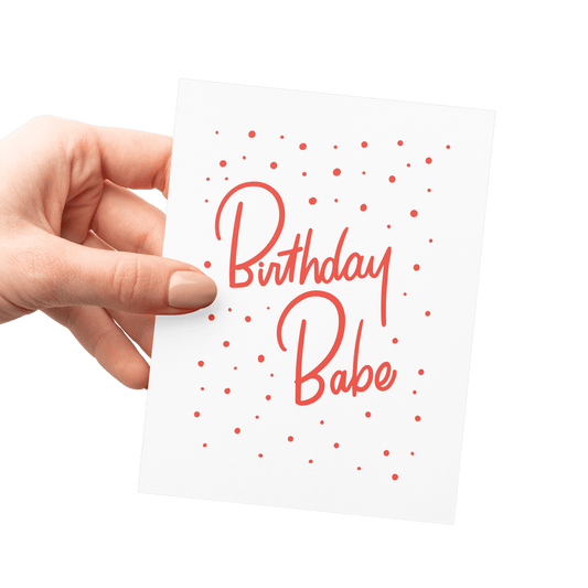 Hand holding a fun 'Birthday Babe' card with red hand lettering and festive confetti design.