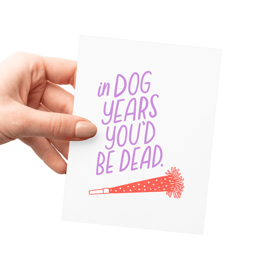 Hand holding a humorous birthday card with 'In dog years, you’d be dead' in lavender hand lettering and a red party horn illustration.