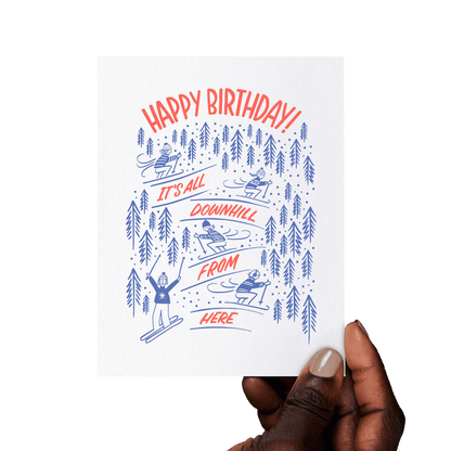 Hand holding a ski-themed birthday card with 'It’s All Downhill From Here!' and playful skiing illustrations.