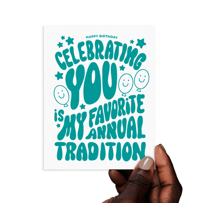 Hand holding birthday card with 'Celebrating you is my favorite annual tradition' in playful green lettering.