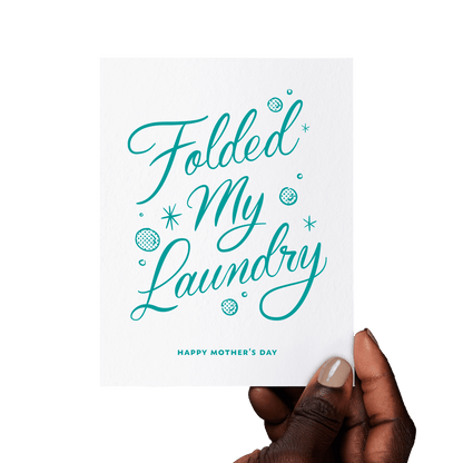 Hand holding a funny Mother’s Day card with ‘Folded My Laundry’ in teal hand lettering, accented with vintage-style bubbles and stars.
