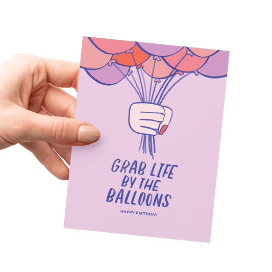 Hand holding a funny birthday card with 'Grab Life by the Balloons' and an illustrated hand gripping balloons.