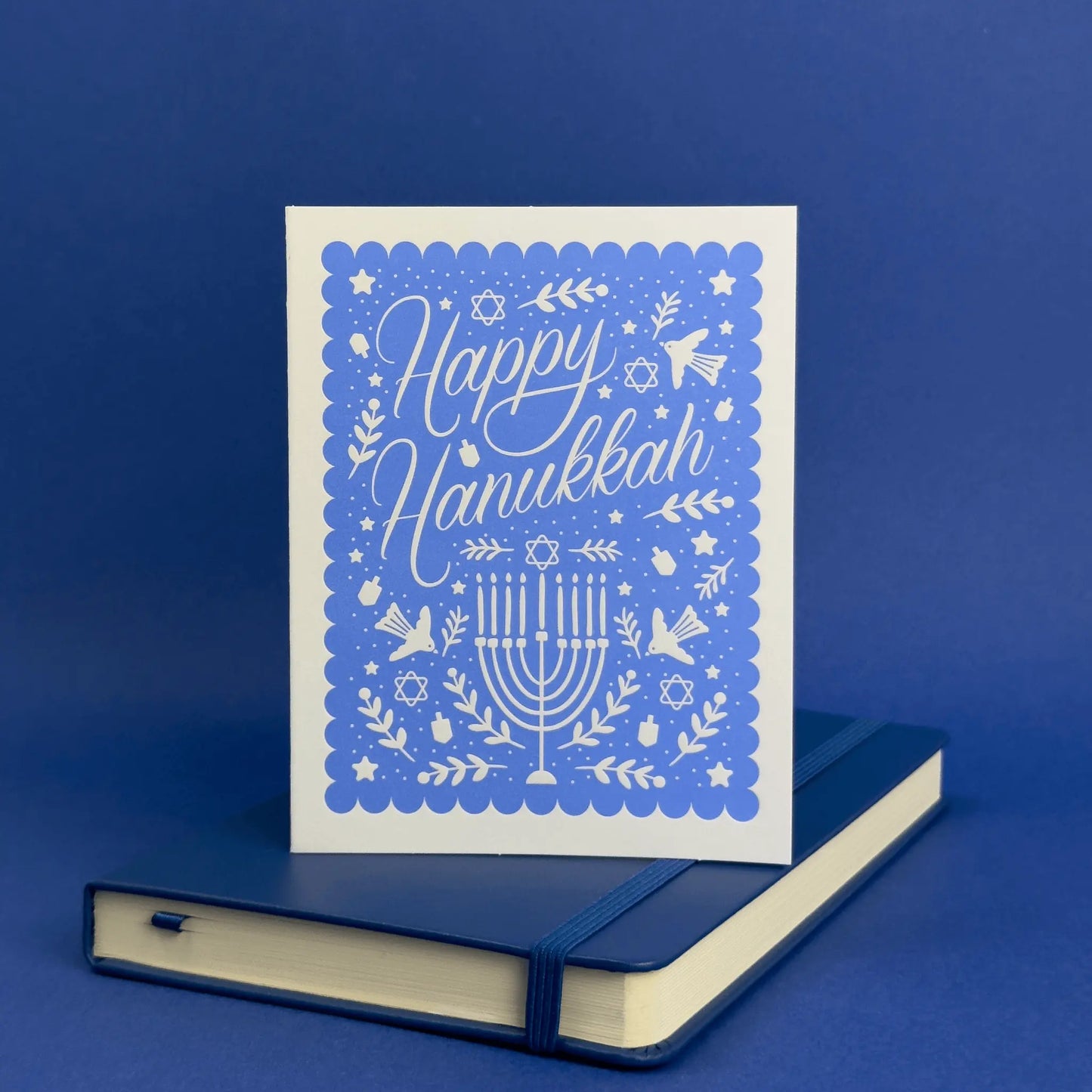 Card with 'Happy Hanukkah' text and menorah design on a blue background, placed on a matching blue notebook.