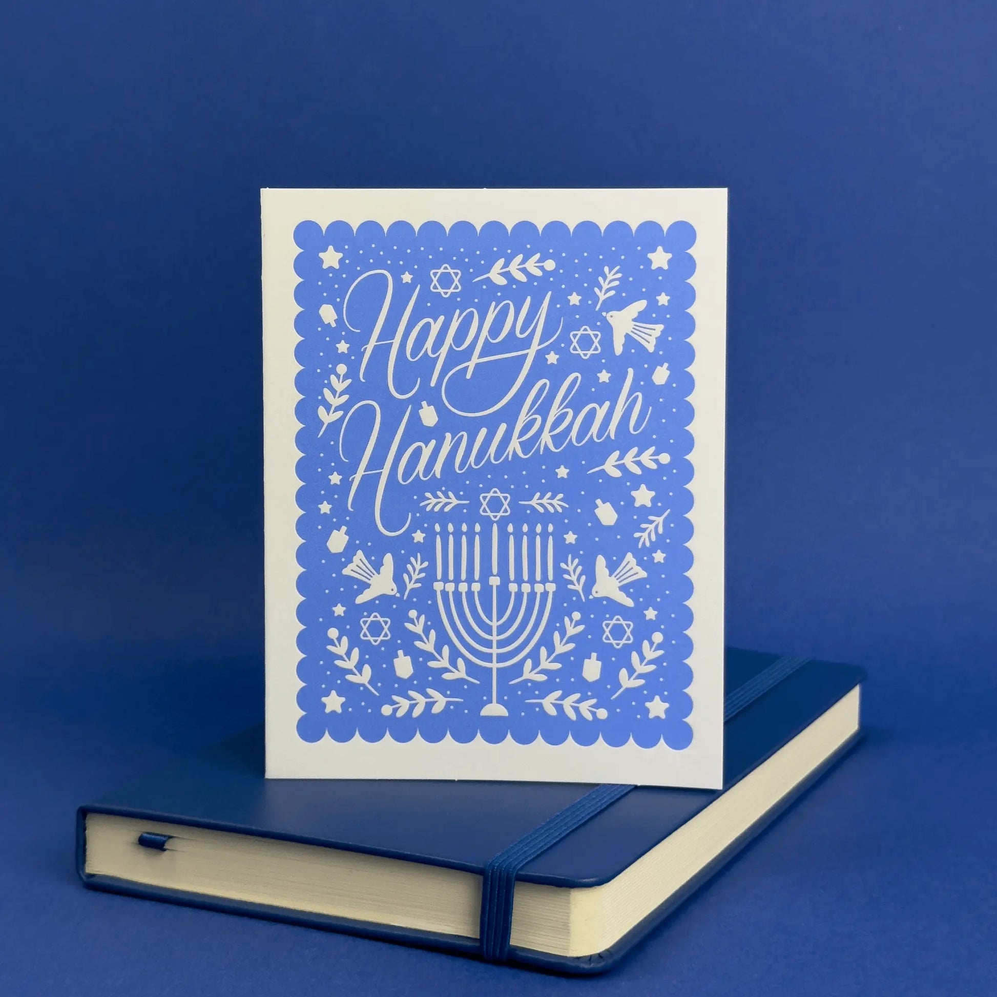 Card with 'Happy Hanukkah' text and menorah design on a blue background, placed on a matching blue notebook.