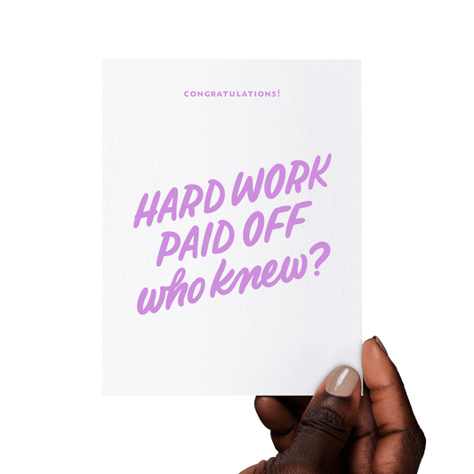Hand holding a funny congratulations card with ‘Hard work paid off—who knew?’ in lavender hand lettering on white cotton paper.
