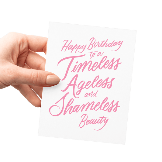 Hand holding a humorous birthday card with 'Happy Birthday to a Timeless, Ageless, and Shameless Beauty' in pink hand lettering.