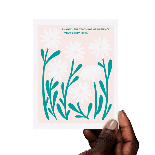 Hand holding a floral greeting card with ‘Thanks for teaching me patience—yours, not mine.’ in teal hand lettering, framed by white daisies on a pink background.