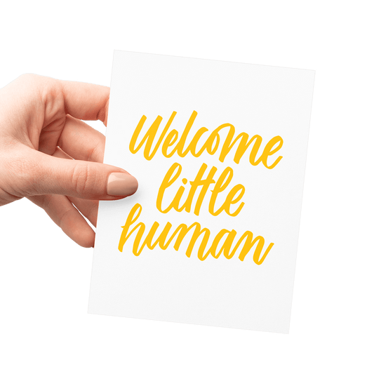 Hand holding a new baby card with 'Welcome Little Human' in yellow hand lettering on premium letterpress paper.