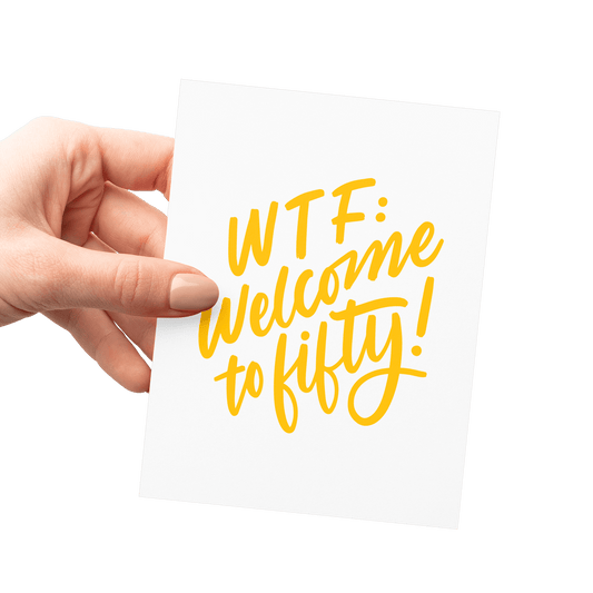 Hand holding a humorous 50th birthday card with 'WTF: Welcome to Fifty!' in yellow hand lettering.