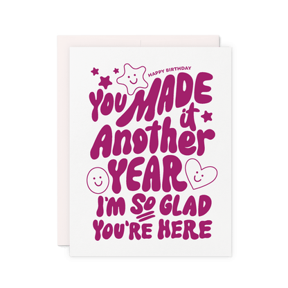 Birthday card that reads 'You made it another year. I'm so glad you're here' in purple hand lettering with stars, hearts, and smiley faces.