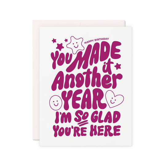 Birthday card that reads 'You made it another year. I'm so glad you're here' in purple hand lettering with stars, hearts, and smiley faces.