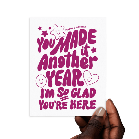 Hand holding birthday card with 'You made it another year. I'm so glad you're here' in playful purple lettering.